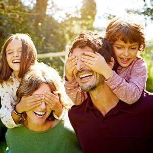 Familia de cuatro disfrutando de un día soleado al aire libre. Dos adultos llevan a dos niños sobre sus hombros, rodeados de árboles y vegetación