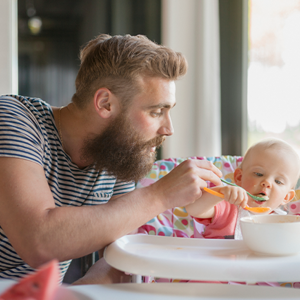 Hombre con barba alimentando papilla a un bebé en una silla alta. En la mesa hay un bol blanco.