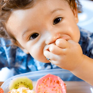 Niño pequeño sentado en silla alta comiendo frutas con las manos.