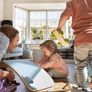 Mujer adulta y dos niños juegan en una sala, con un tablero de equilibrio y un coche de juguete.