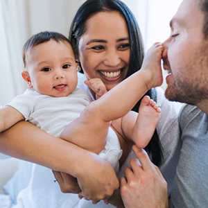 Mamá carga al bebé sonriendo. Bebé tiene el pie en la nariz del papá.